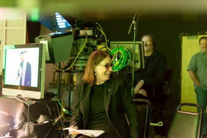 A photo of a white, brown-haired woman wearing glasses and a black blazer and black t-shirt in front of a camera on a TV set.