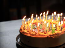 A single-layer, round birthday cake with many lit candles is shown in close-up. 
