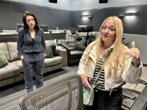 Two female presenting persons stand inside a room painted gray. Several couches are behind them.