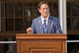 A white man with short, dark brown hair stands at a lectern. He's wearing a blue suit, white shirt and blue diagonally striped tie. 