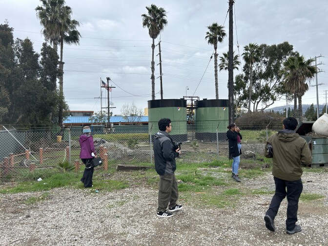 A group of four people walk next to large green cylindrical oil holding tanks in a gravel and grassy lot. 