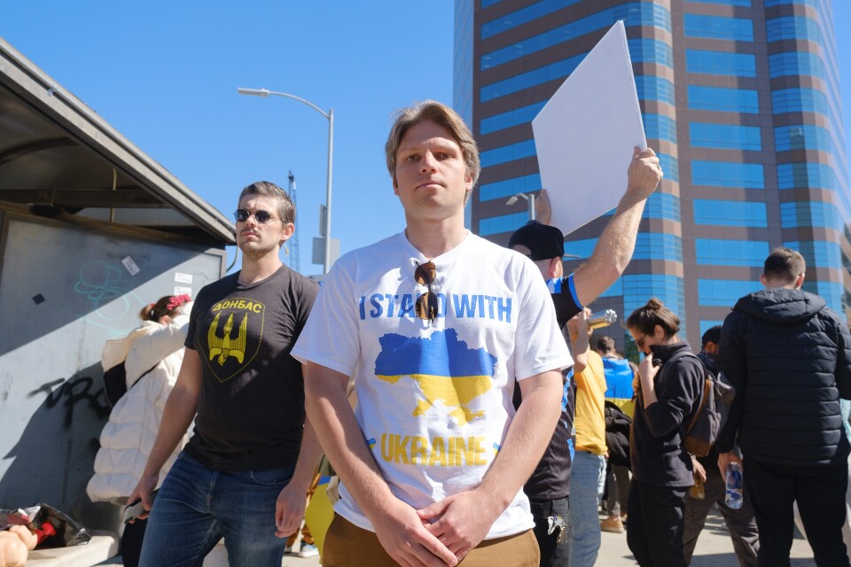 A male protester holds a microphone and addresses the crowd. He is wearing a t-shirt with a slogan that reads: "I Stand With Ukraine."