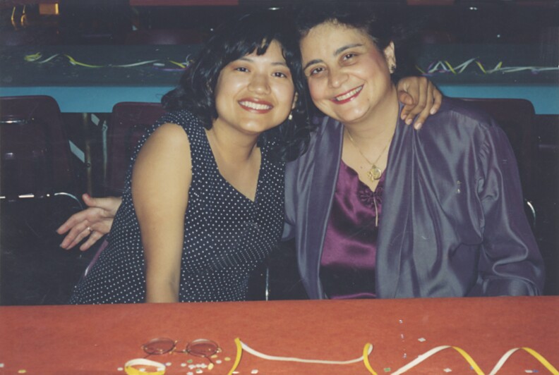 Two women embrace each other and smile while sitting at a long table.