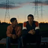 Lamar Johnson and Aaron Pierre sit on a picnic table in a park at dusk, the small buildings and cell phone towers just silhouettes behind them. Johnson sits on the left, he wears jeans and a tan leather jacket. His elbows rest on his legs, his hands clasping each other in his lap. Johnson looks over at Pierre who sits in the exact same position wearing black pants and a red, blue, and white color blocked jacket. Pierre looks at the ground not meeting Johnson's gaze. 