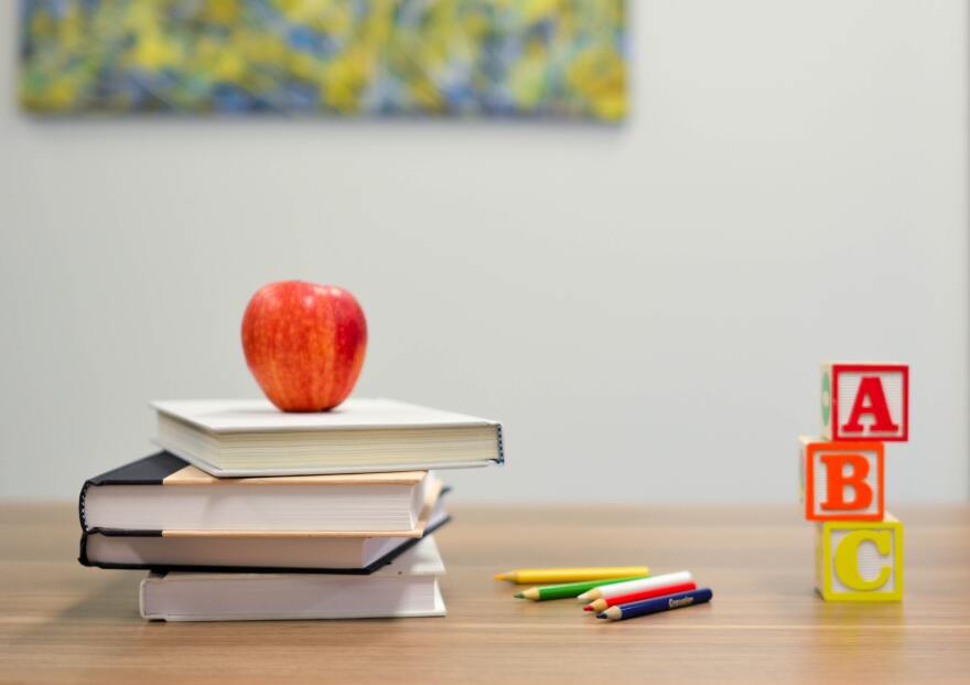 Four hardbound books stacked on a desk, with red apple resting on top. A handful of colored pencils lie nearby, and letter blocks are stacked to the right of those. The blocks spell "A-B-C."