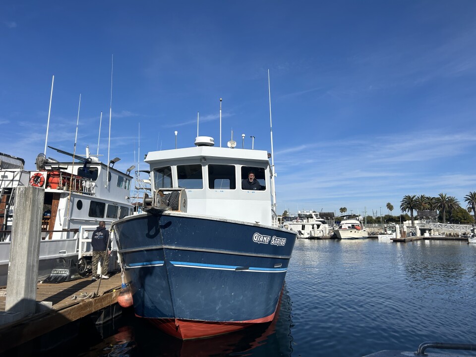 An image of a navy blue fishing boat under blue skies and calm blue water in a harbor. 