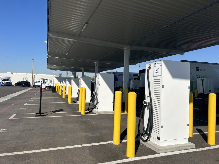 White rectangular boxes that are heavy-duty truck charging stations lined up next to each other in a parking lot on a sunny day. 