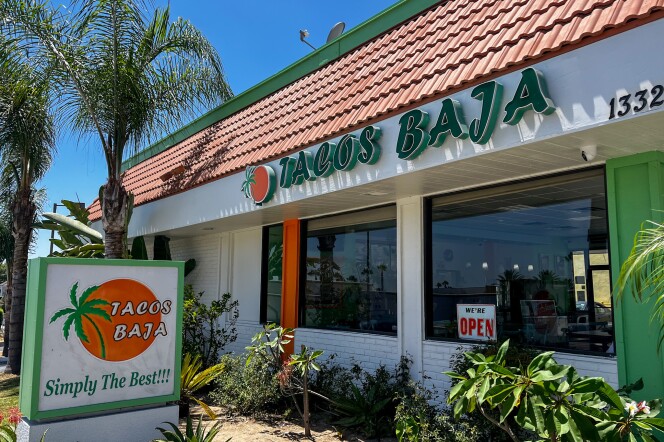 The building exterior features a slanted roof with green lettering signage that reads "Tacos Baja." Below the sign, there are two windows—one with an open sign. The building is surrounded by lush green foliage. In the foreground, there is a standalone sign that reads "Tacos Baja, Simply The Best."