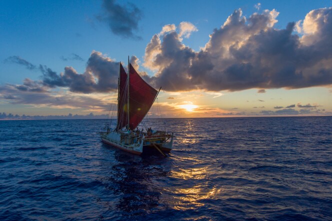 The Hōkūleʻa, a replica of an ancient two-hulled Polynesian voyaging canoe, sails into the sunset under blue skies and high fluffy clouds. 