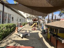 A long playground is next to a one-story white building to its left. The playground has a sand floor, a wooden structure with a slide and various other equipment. To the right is another wooden structure, also on the sand. It's a sunny day and blue sky can be seen at the top of the frame. A green hedge lines the white building.