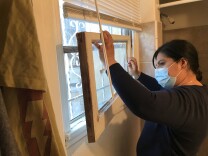 A woman wearing a mask in her apartment bathroom removes a broken window from its frame.