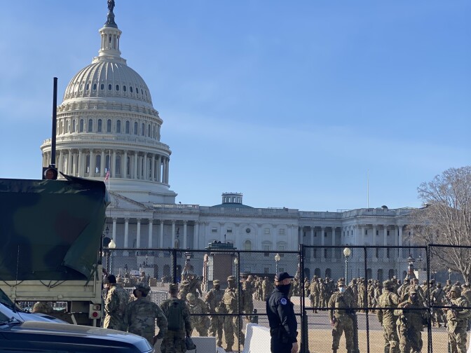 The U.S. Capitol building is peaking out in the distance behind large, black, non-scalable fences. Scores of soldiers in uniform are walking around behind the fences, in front of the building. 