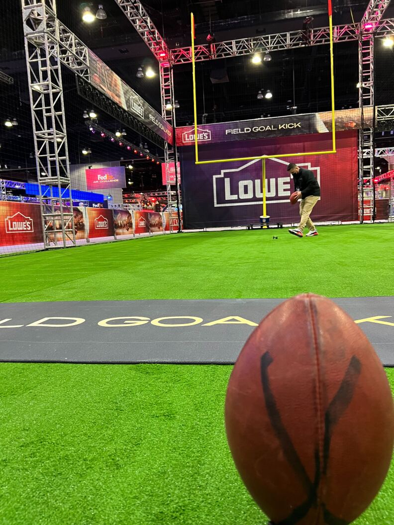 A football is place in on the floor of a green artificial turf surface in the foreground with a yellow goalpost in the background at an activity where visitors to the Super Bowl Experience can practice their kicking skills.