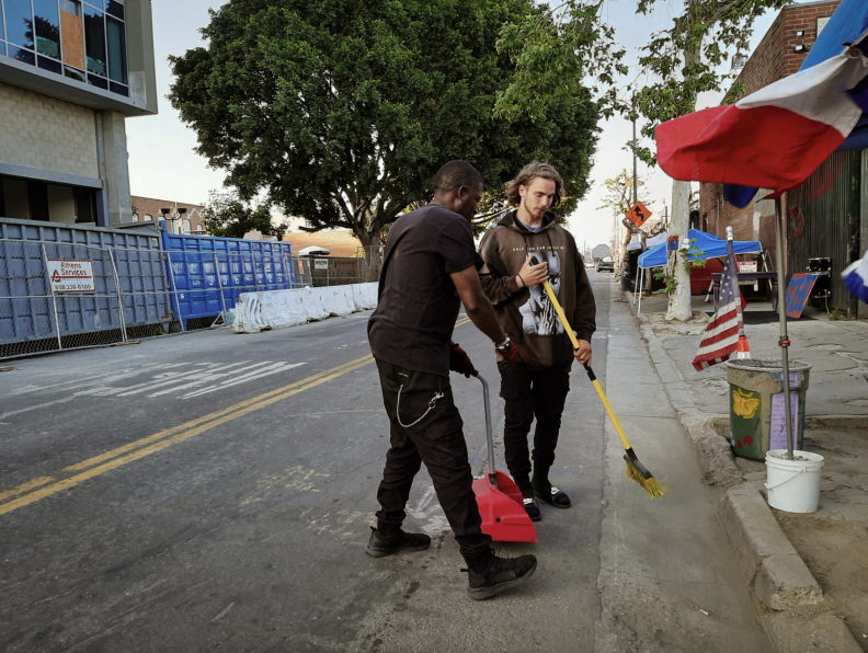 A man with dark brown skin tone and another man with light skin tone and long hair sweep streets during the daytime.