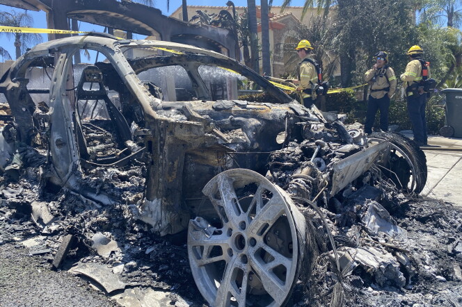 The burned out husk of a car, wheels left with no tire around them, ashen pieces/remnants all around, as firefighters stand nearby. Yellow caution tape extends in the background, with an intact house behind as well.