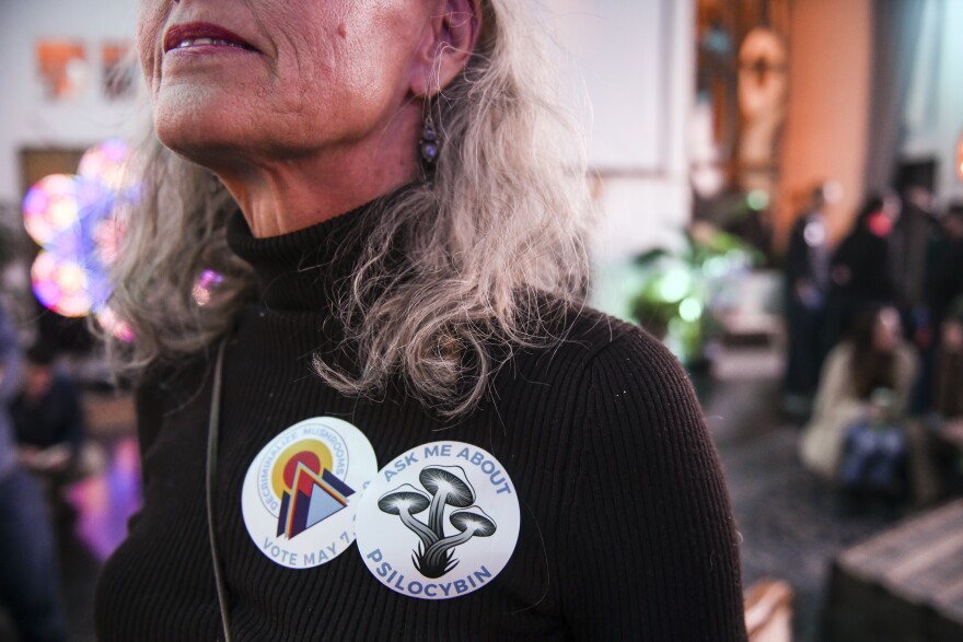 A close-up of two stickers on the shirt of a woman who appears to be white. The stickers are round, and one reads "ask me about psilocybin" and the other reads "vote may 7." The woman's shirt is black and her hair is gray and shoulder-length.