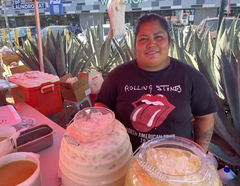 A woman stands behind a table with two large buckets of beverages intended to serve customers.