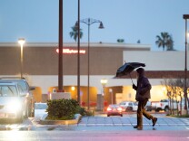A man wearing black pants and black jackets, holds a black umbrella while walking down a side walk in front of a parking lot with a CVS Pharmacy.
