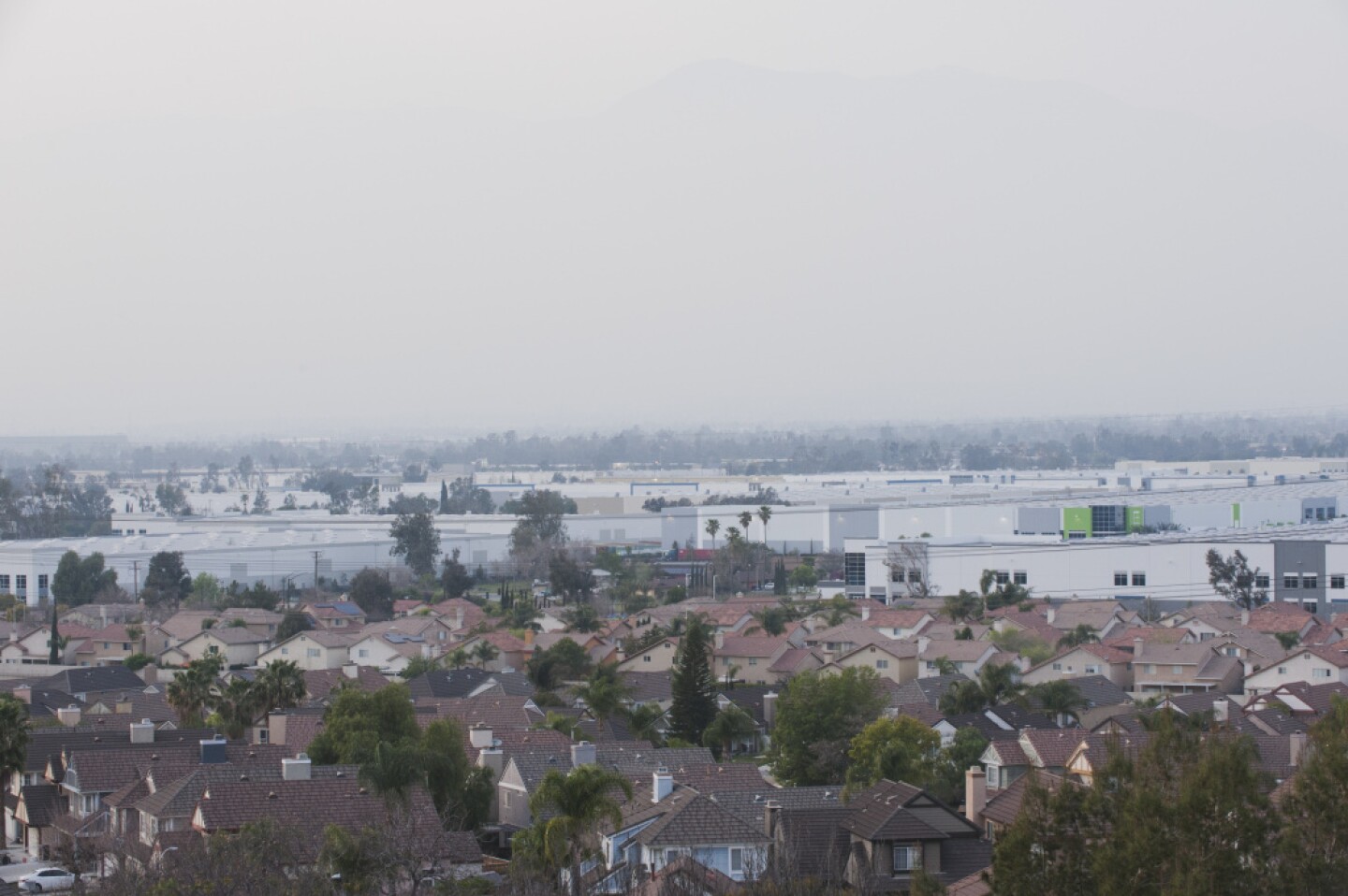 A view from above shows tract homes in Fontana, California, in the foreground and rows of white warehouse behind them.