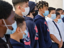 Adolescent boys, most of whom are Black or Latino, stand in a line with face masks, school sweatshirts or white button-downs and ties. They are shown from the side. 