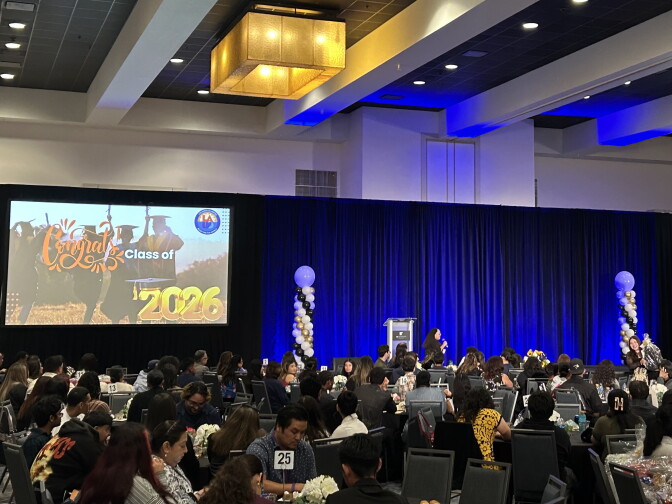 A hotel ballroom filled with dozens of dinner tables and a stage lined with bright blue curtains. A screen is set up on the left of the stage with an image that reads "Congrats Class of 2026." A pair of white, black and gold balloon pillars are set up on either side of a silver podium toward the center of the stage.