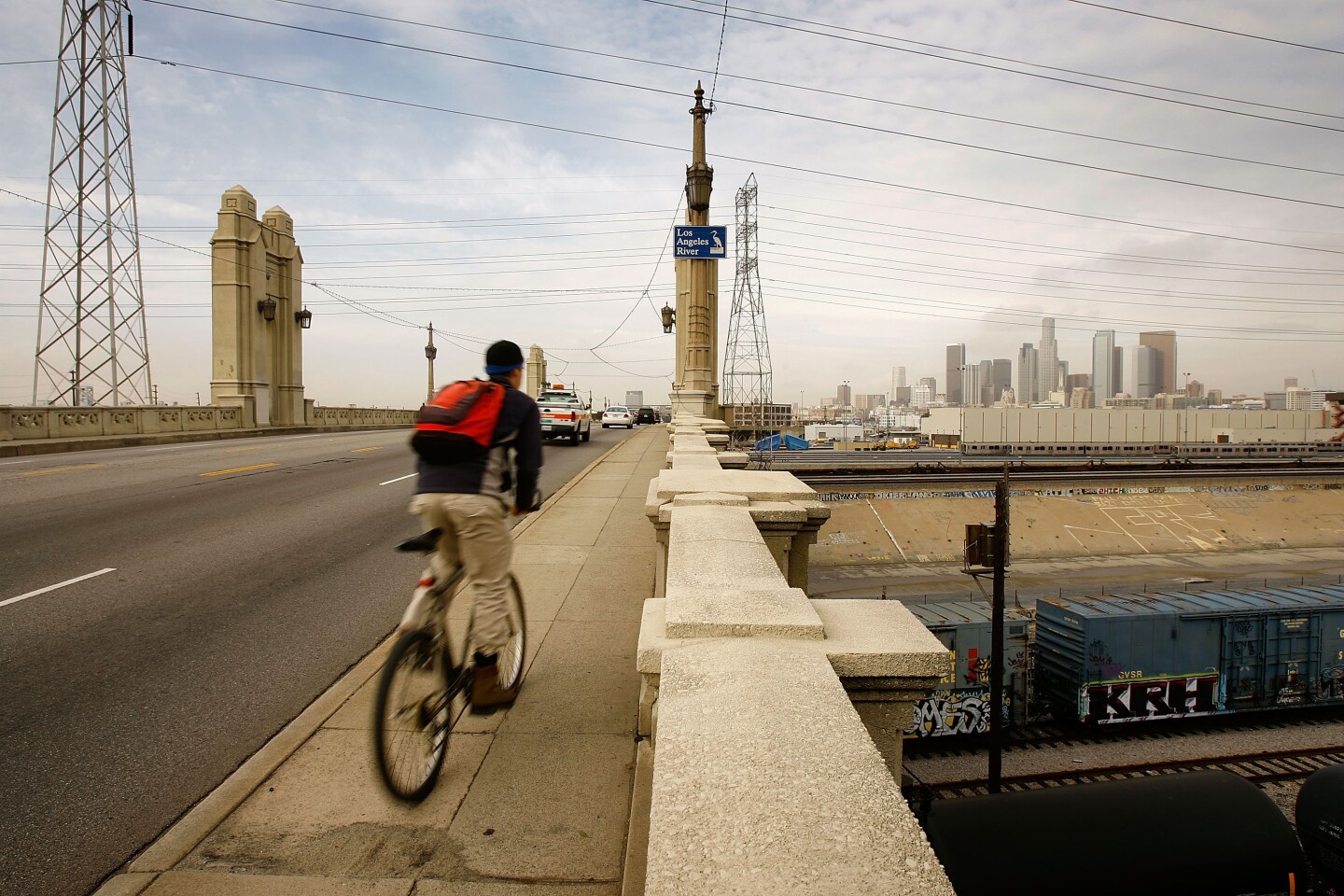 An image of a man wearing a red backpack rides a bicycle across a bridge