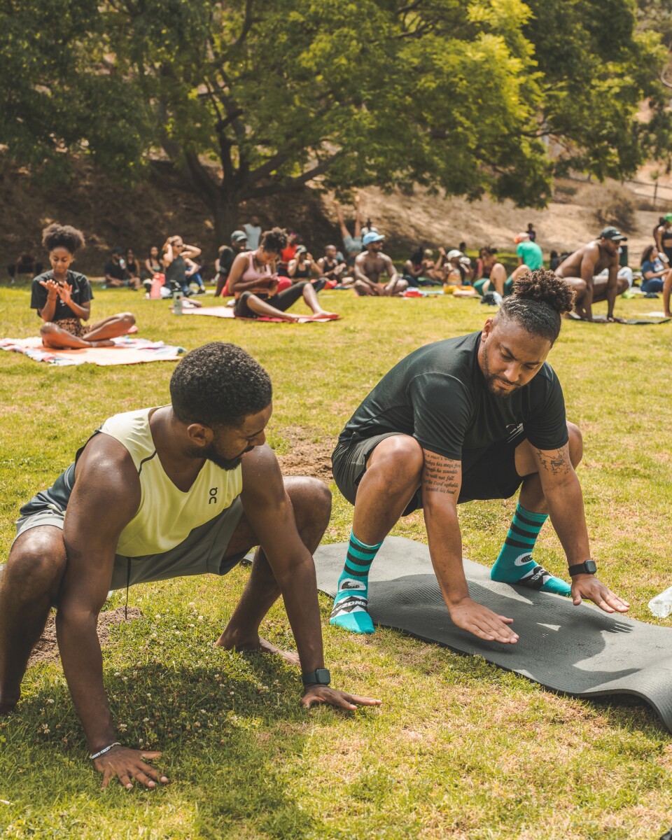 Two brown-skinned Black men engage in a yoga pose at a park. 