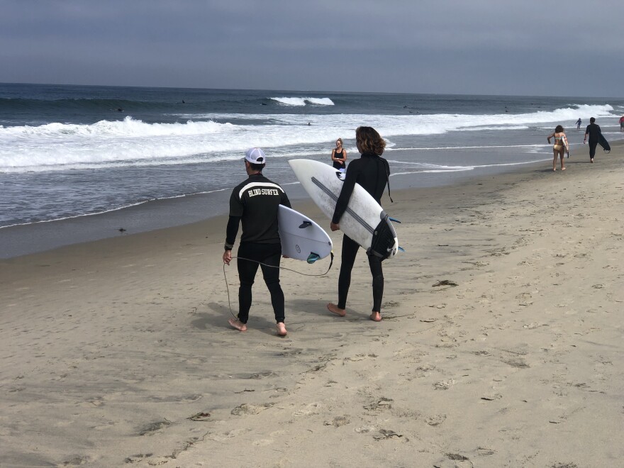 An image of voiceover artist Pete Gustin, who is blind, holding a surfboard under his right arm as he walks on the beach towards the ocean. He wears a t-shirt over his wet suit that reads on the back: BLIND SURFER. He is accompanied by his guide, who also holds a surfboard.