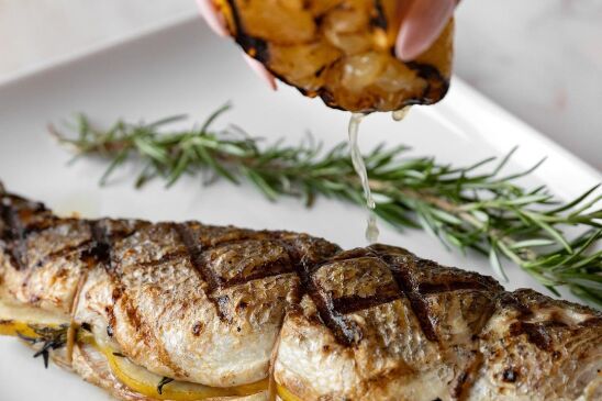 A cooked branzino lies on a white plate, next to a sprig of rosemary. Above it, a manicured hand is squeezing lemon juice onto the fish.