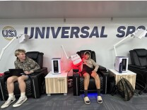 Two adolescent boys sit in black cushioned reclining chairs pushed against a wall displaying the logo USI. Red light therapy machines stick up from consoles between the chairs, and one of the boys has his arm under the red light. 