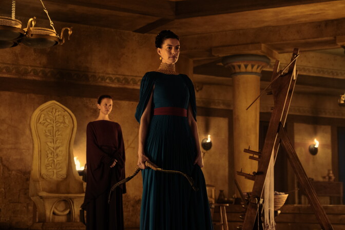 Mia Goth and Anne Hathaway are dressed in dark, ancient clothing inside a warm-toned Greek room.