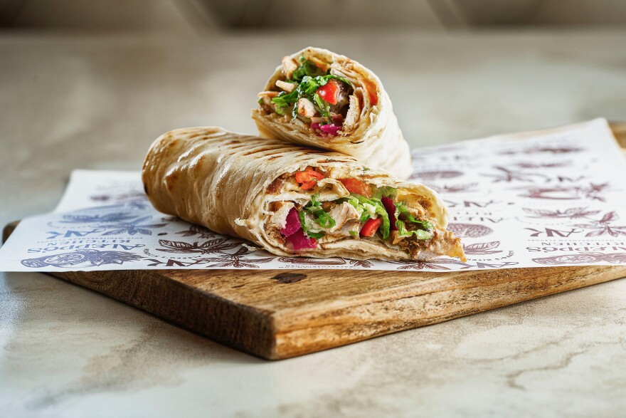 A pita wrap with green lettuce, grilled chicken, pink pickles, red tomato slices sits on a paper on a wooden cutting board.