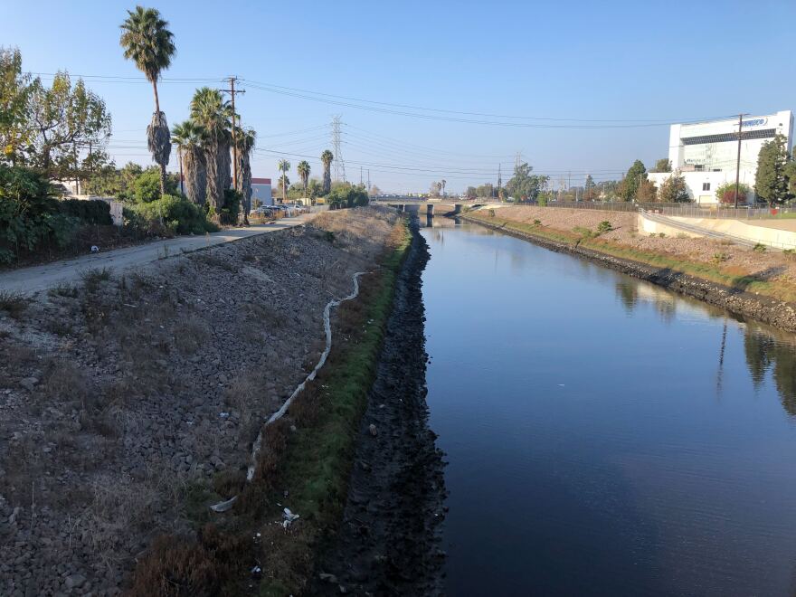 A channel of water is flanked by dirt walls on both sides. A concrete path sits to the left and white building sits to the right. In the distance there is an overpass above the channel.