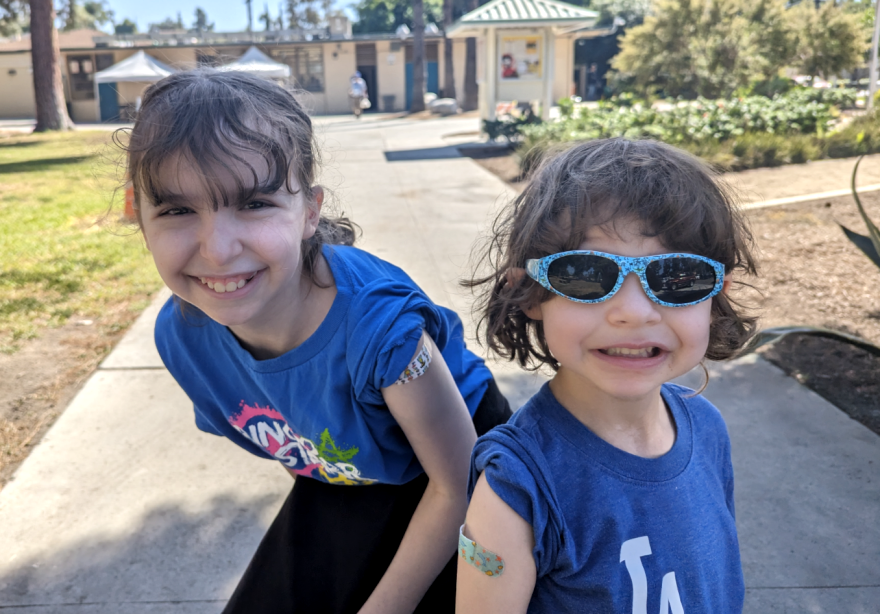Two children outside smile at the camera, both with bandaids on their arms after getting vaccinated.