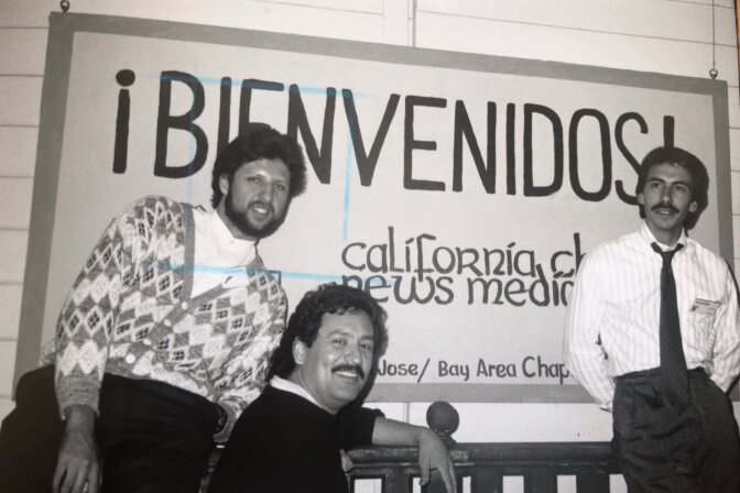 Black and white photo of three male-presenting people with mustaches, in front of a sign that says welcome in Spanish.