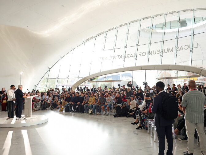 A speaker addresses a seated crowd and photographers inside a glass-walled hall reading 'Lucas Museum of Narrative Art.'
