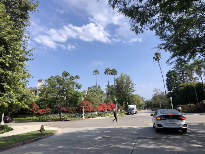 A white hybrid vehicle on a shady street, lined with trees and flowers, on a sunny day with blue skies. A woman walks across the street in front of the vehicle. 