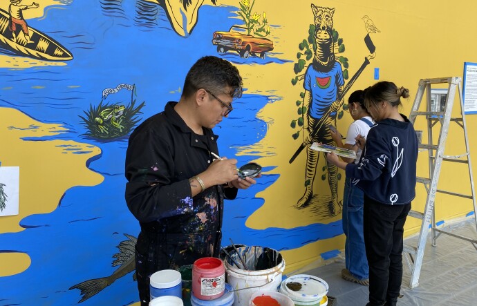 Three people work on a mural, which features a body of water and other elements, including a fisherman casting a net and a lowrider sprouting flowers. There are painting tools around the three people, including a ladder and multiple buckets of paint. The floor has been sealed off with plastic and blue tape. 