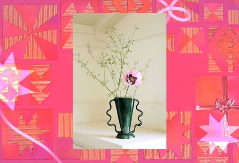 A green, glass vase holds a purple flower in bloom, and sprigs of greenery. The vase is also accented with a wavy ribbon design. The image is set against a festive turquoise background.