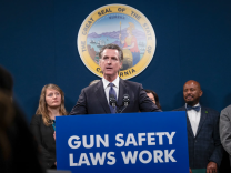 Governor Gavin Newsom, a light-skinned man with salt-and-pepper hair wearing a charcoal gray suit and blue tie, stands at a podium labeled GUN SAFETY LAWS WORK in white lettering on a blue background. The California state seal is posted behind him. Four people stand behind him a the podium.