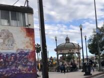 To the left a wall has a mural of a woman with graffiti over some landscape. To the right a park square features an elaborate  gazebo. People are walking through the park.
