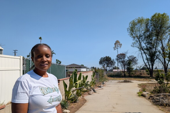 Woman with short black hair wearing white t-shirt with 'FORWARD EVER' text stands on concrete path surrounded by desert plants and trees under clear blue sky.