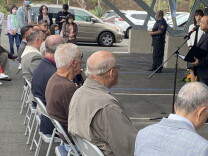 Reverend Albany Lee leads members of the Laguna Woods and larger Orange County community in prayer. He stands in front of bouquets of flowers. Members of the community sit on folding chairs that have been set up in the church parking lot. 