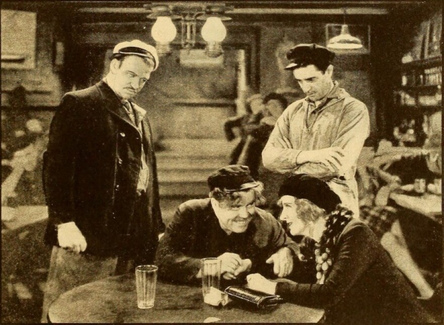 Two men in sailors caps stand over another man and a woman sitting at a table in a bar.