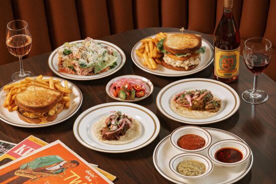 A table set with a wide variety of dieshes, from a patty melt with fries to tacos. There's also a bottle of wine on the table and two full  glasses. 