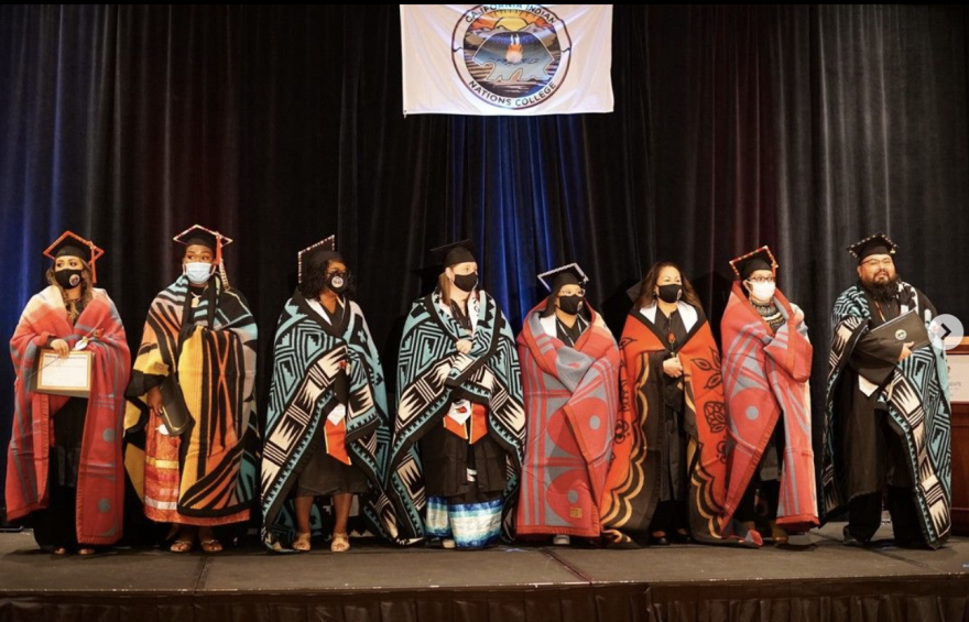 Eight men and women wearing graduation caps, face masks and wrapped in colorful blankets stand next to each other on stage. Above and behind them hangs a banner that reads California Indian Nations College.