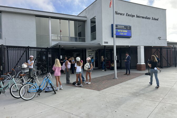 In front of a school building, a group of students stand outside. Three bicycles are parked out front. 
