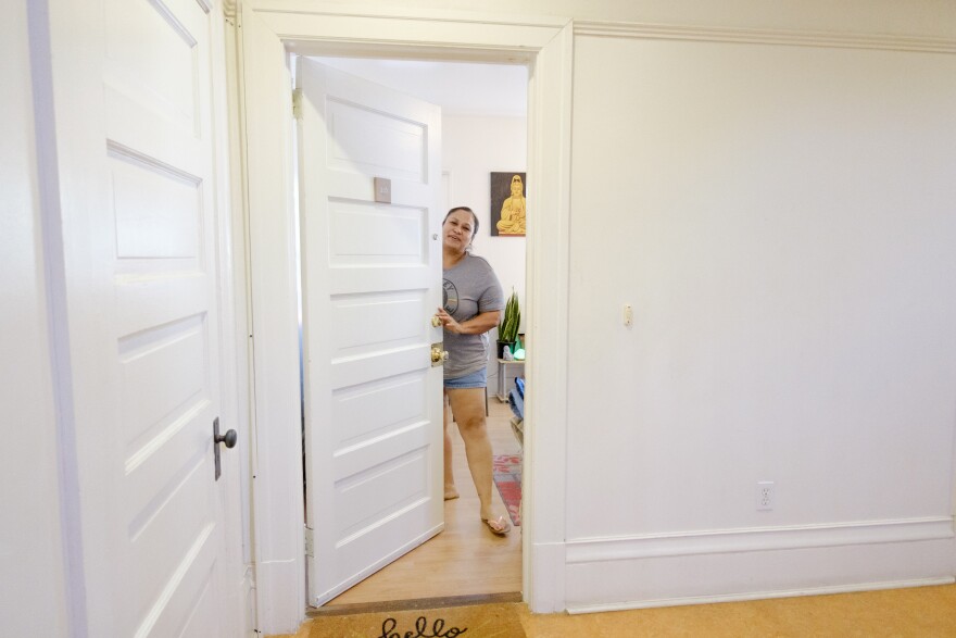 A formerly unhoused woman stands inside her apartment while holding the front door open. She is wearing a gray t-shirt, blue jean shorts and sandals.