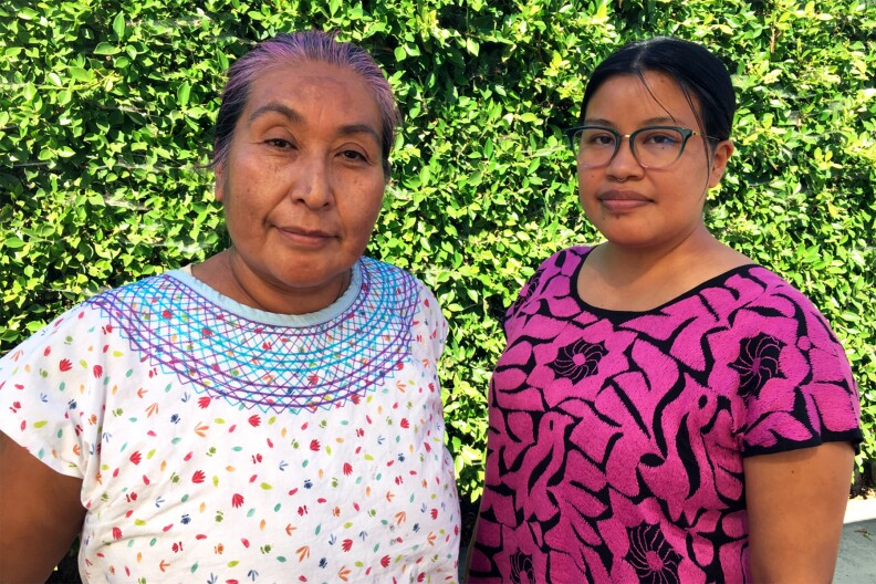 Two brown-skinned women against a background of green foliage; one on the left is in a white printed blouse, the other on the right in pink.