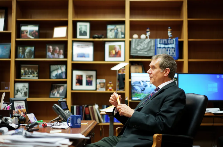 L.A. County Superior Court Judge Ronald Owen Kaye, a man with light skin tone, wearing a dark suit, looks to the side as he sits in a chair behind a desk in an office. There are framed photos sitting on shelves in the background.
